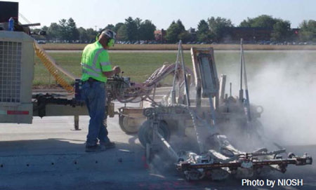 2015-200Fig1 Vista posterior de un trabajador parado con las piernas cruzadas y con protección auditiva puesta. Sus brazos están doblados y sus manos se encuentran sobre los controles de la máquina de perforación para pasadores.