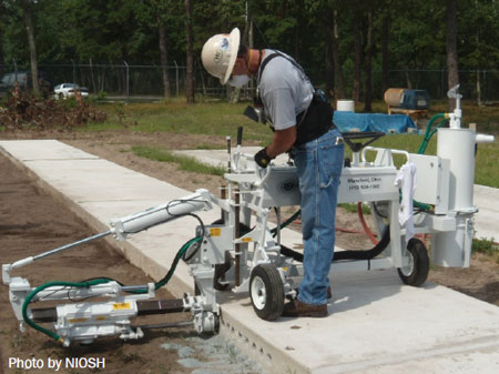 2015-200Fig2 Vista lateral de un trabajador inclinado hacia adelante, de la cintura para arriba, usando un casco y máscara de protección contra el polvo mientras está parado en un tramo de concreto y opera una máquina de perforación para pasadores.
