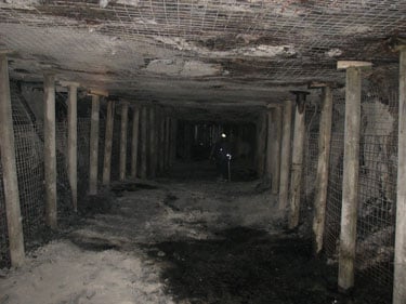 Fotografía que muestra postes de madera alineados cada lado del pasaje de una mina subterránea y que sostienen el techo. Se muestra malla soldada contra la pared y en el techo para que proteja contra los derrumbes del techo y los pilares de seguridad.