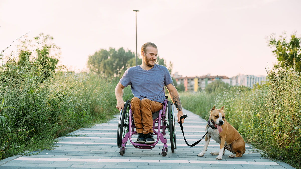 Man in a wheelchair on a paved path smiling at his dog sitting beside him outdoors