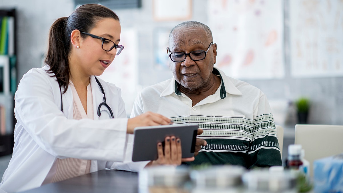White female doctor with brown hair in a low ponytail with Black older adult patient looking at a tablet.