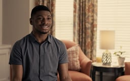 Black male sitting in room with window and chair behind him talking to camera