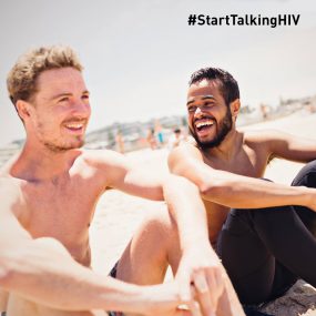 Image displays two men smiling/laughing while sitting on a beach.