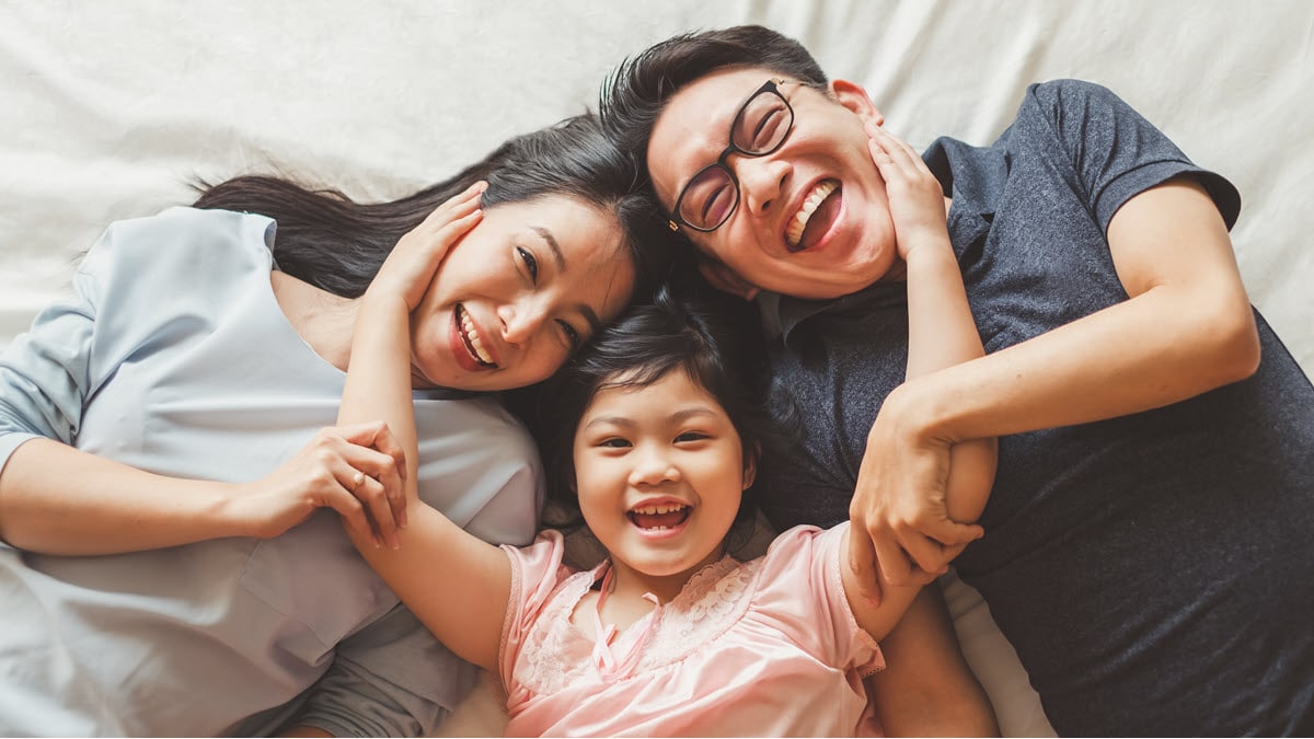 A happy Asian family laying on a bed laughing with parents and daughter in the middle hugging them