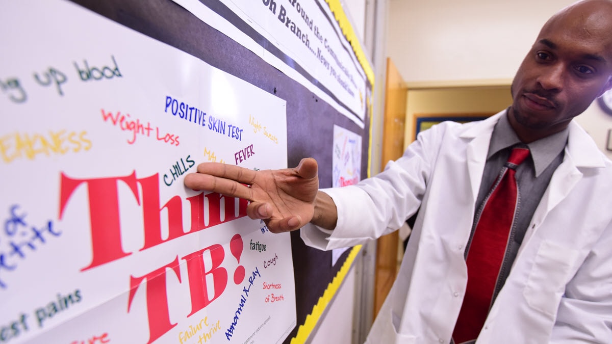 A health care provider points to a poster with "Think TB!" printed in red.