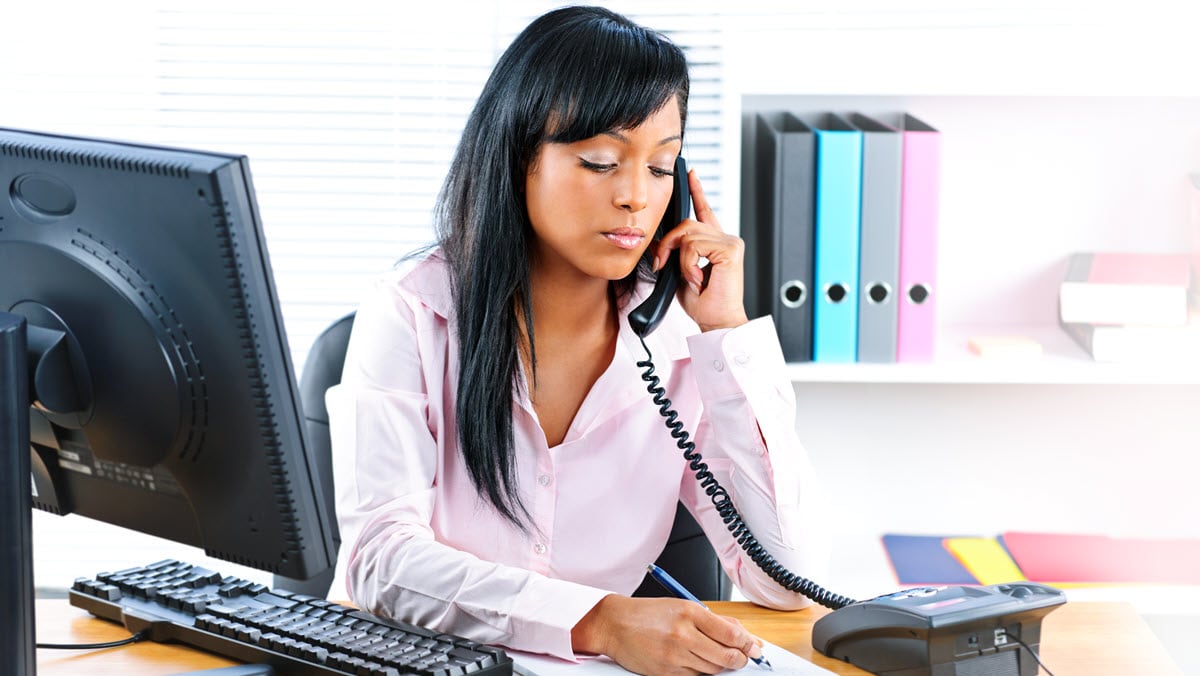 A woman seated at a desk listens on a telephone while taking notes.