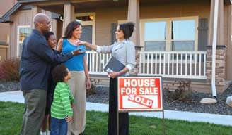 esposa, esposo y dos hijos hablando con una agente inmobiliaria delante de una casa con cartel indicando que está vendida.