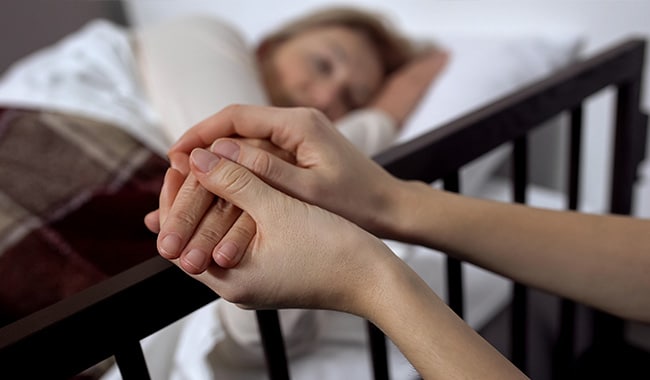 Caregiver cradling the hand of a loved one lying in a hospital bed.