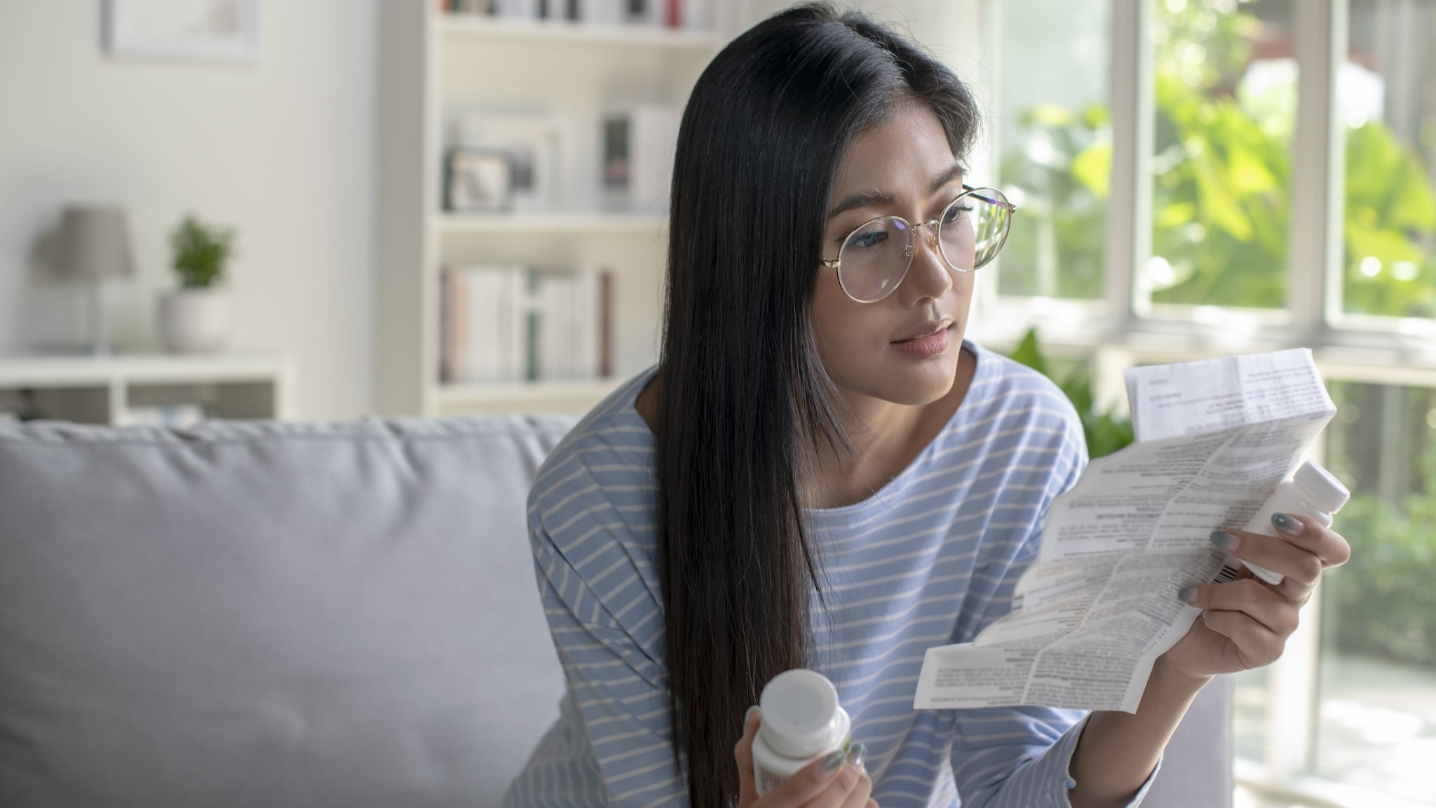 An adult with long hair and glasses is reading an information sheet in their left hand for a medication bottle in their right hand.