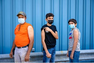 A latino man and two latino boys in masks showing their bandaids from where they were vaccinated.
