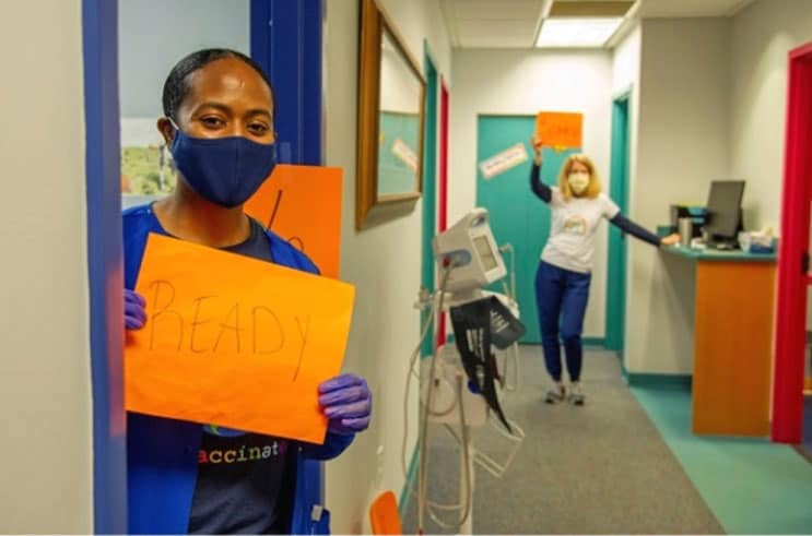 COVID-19 vaccine: DeKalb Pediatric Center’s medical assistant and doctor hold signs to indicate they are ready for the next person.