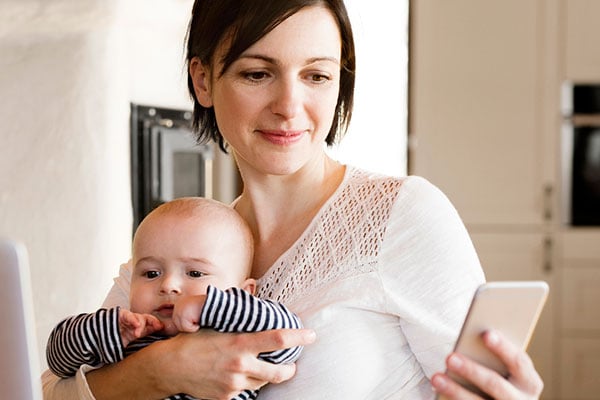 Mother at home with baby working on laptop, holding smartphone