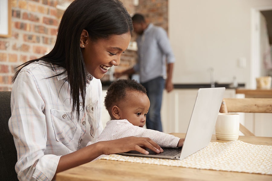 Madre escribiendo en su computadora con su bebé en brazos.