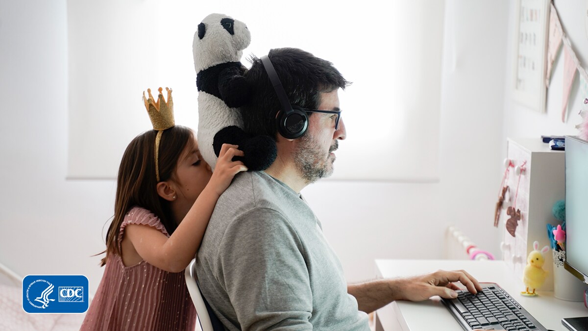 Daughter standing behind her father holding a stuffed panda on his shoulders as he is on the computer.