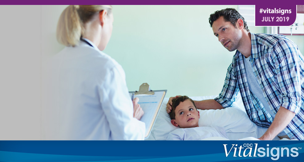 A female doctor talks with a boy laying in a hospital bed who is a patient, and his father.