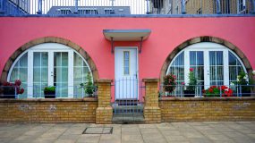 front view of brick building entrance, painted pink, with curved windows on each side and flower pots