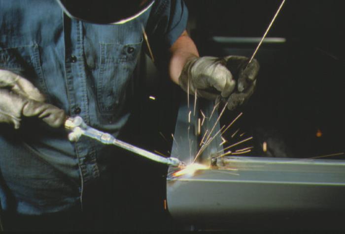 A welder in denim coveralls and a welder's mask works on metal