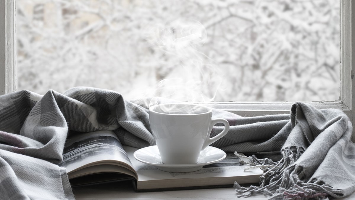 Steaming hot drink in a cup sitting on a book and next to a warm scarf on a snowy day