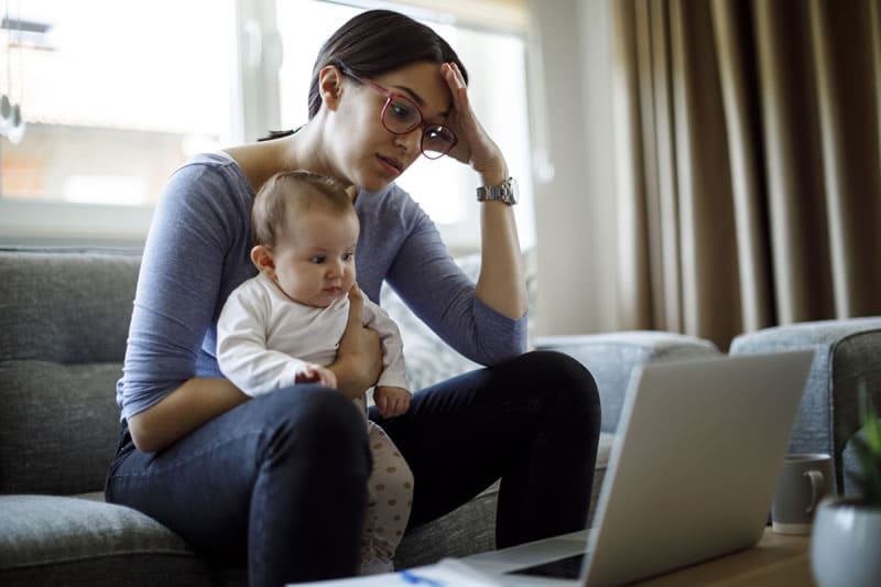 Mujer sentada en un sofá con un bebé en brazos y mirando una computadora portátil