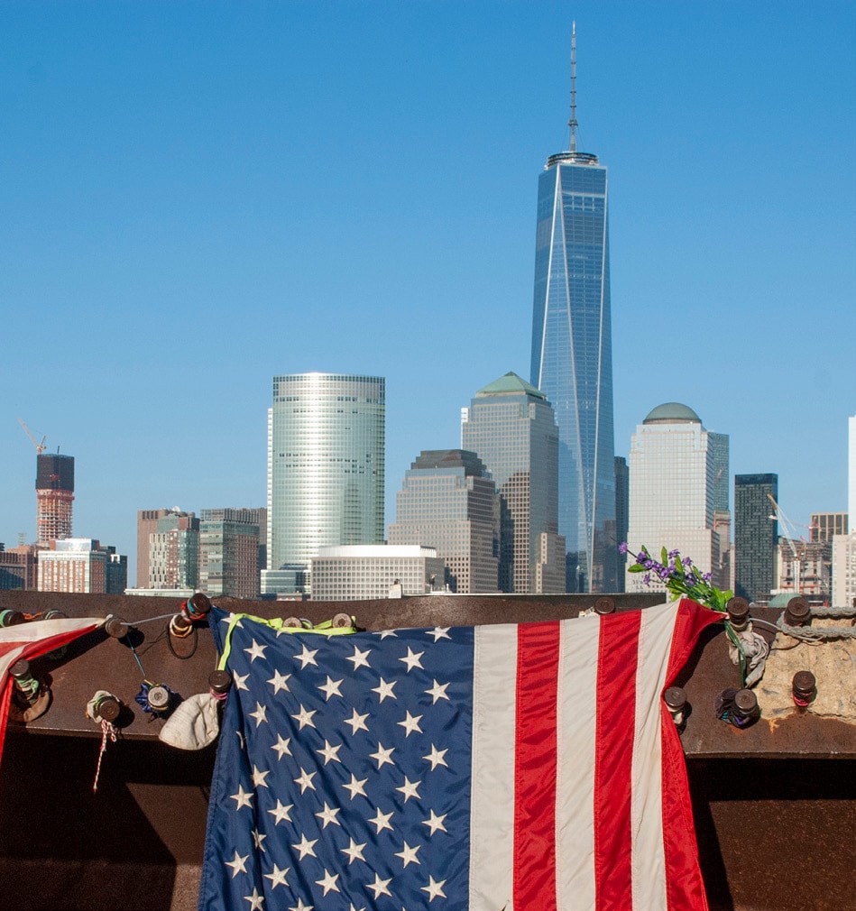 Image shows an American Flag hanging in the foreground of the Lower Manhattan skyline.