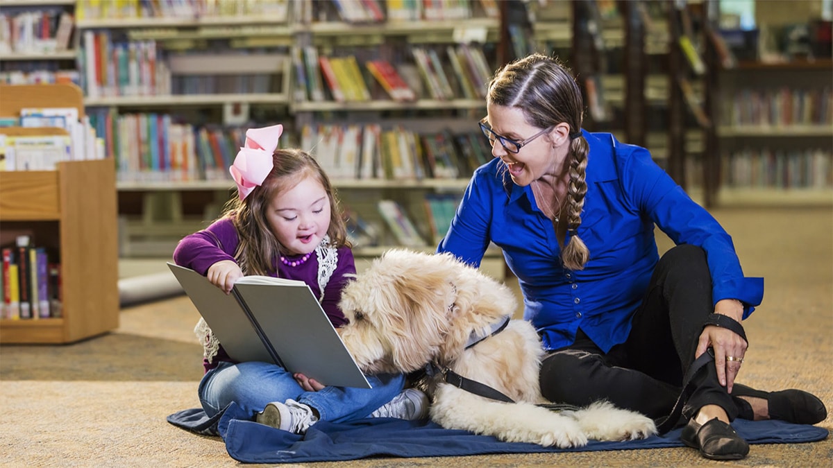 School librarian sitting on the floor reading a book aloud with a young girl and the girl's service dog