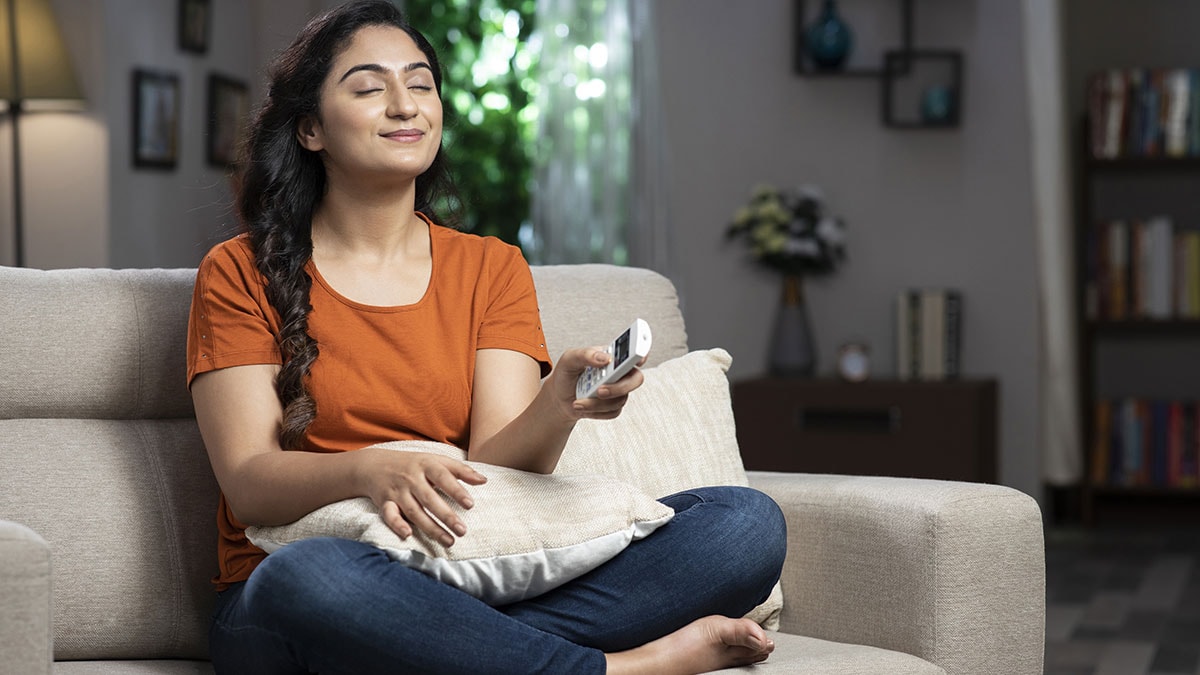 Young woman sitting on a couch and turning on the air conditioning with a remote.