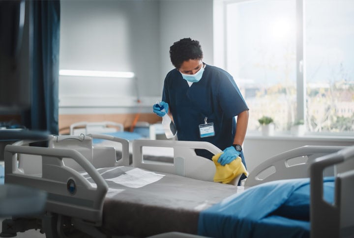 Female healthcare worker wearing surgical mask wiping down hospital bed in patient room. She is holding a spray bottle and cloth.